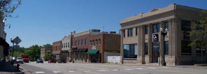 Small-town main street with historic brick storefronts and a neoclassical bank building under a clear blue sky.