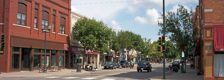 Downtown street lined with historic brick storefronts, trees, and local shops on a sunny day.