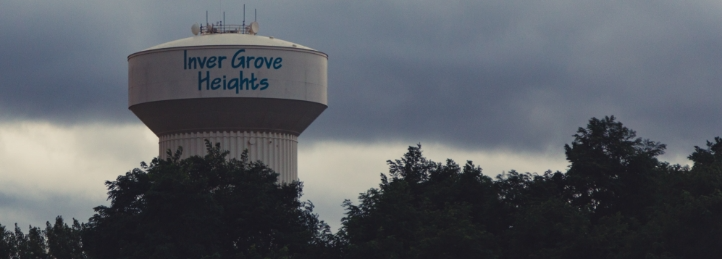 Inver Grove Heights water tower rising above a tree line under dark cloudy skies.