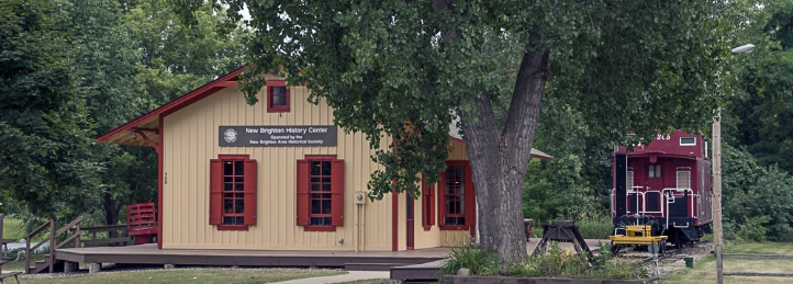 New Brighton History Center building with vintage red caboose and surrounding trees in a park setting.