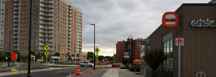 Urban streetscape with modern apartment towers, retail storefronts, and road construction under cloudy skies.