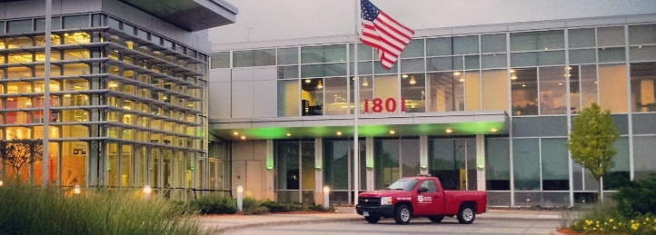 Modern office or civic building entrance with large glass windows, American flag, and red pickup truck parked outside.