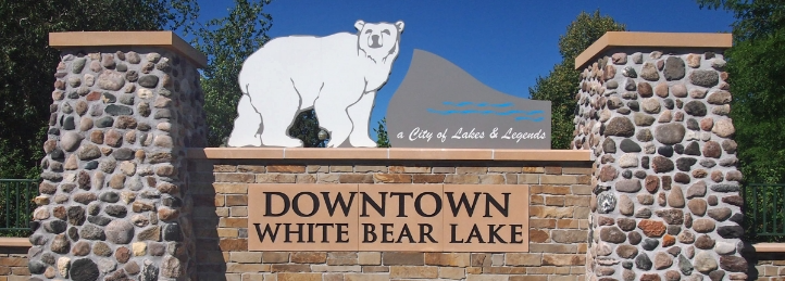 Downtown White Bear Lake entrance sign with stone pillars, polar bear graphic, and clear blue sky.