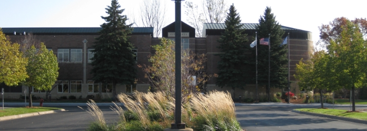 Modern civic or community building with landscaped parking area, evergreen trees, and American flag display.